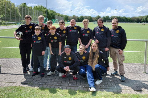 Dreizehn Konfis und drei Betreuer als Gruppe vor einem leeren Fußballfeld.